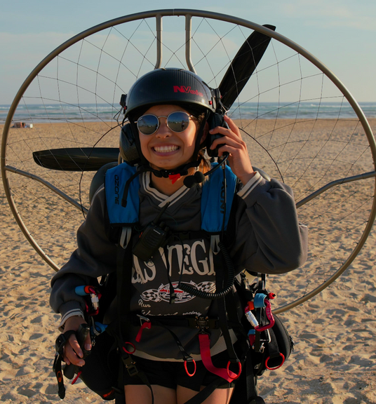 A person on a beach wearing a powered paraglider setup, including a helmet, headset, harness, and wing frame, with the ocean and sand in the background.