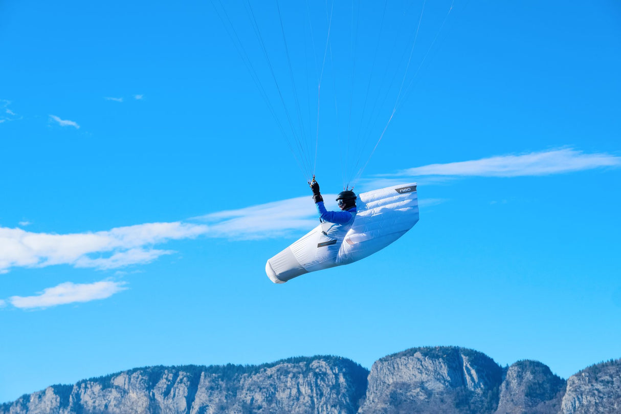 Person in a white hand gliding harness flying against a blue sky with mountains in the background