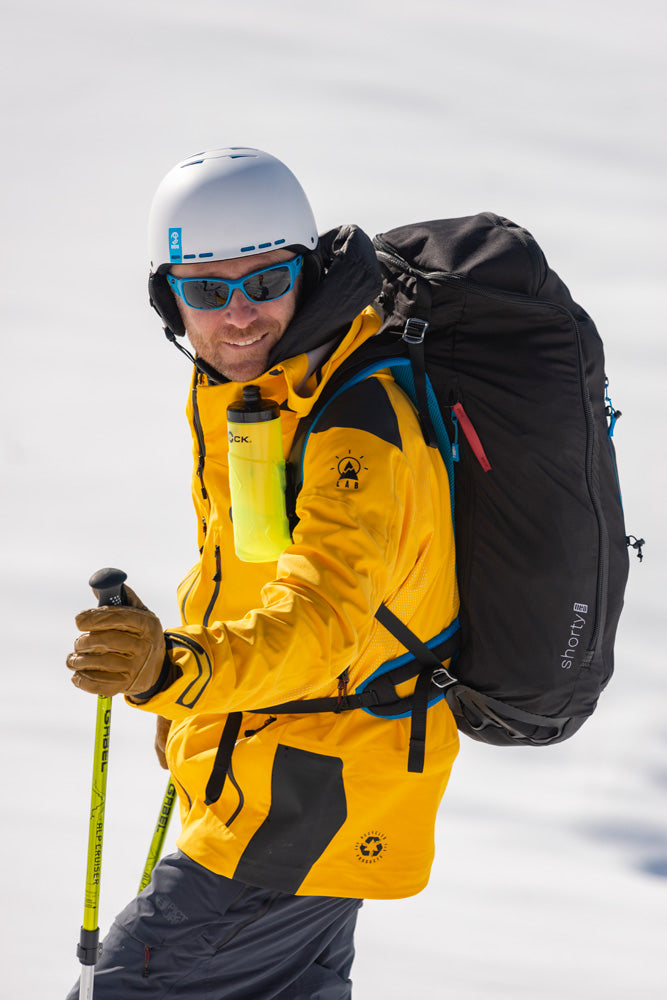 Person in yellow jacket with black NEO shorty paramotor harness and ski equipment on a snowy background