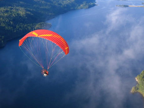 Red and yellow paraglider soaring over a blue lake with green trees.